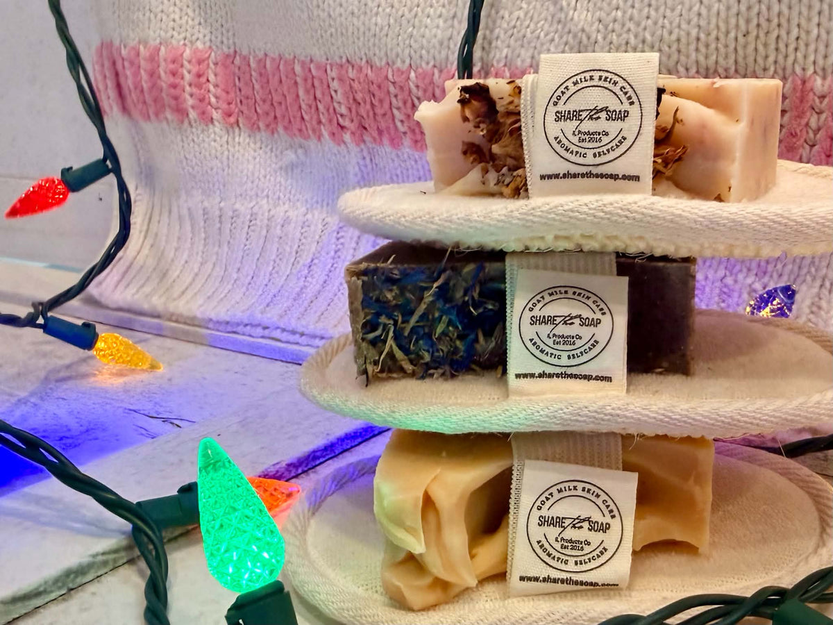 Three handmade artisanal soaps in different colors and textures displayed on a three-tiered white stand, decorated with colorful Christmas lights and a pink knit blanket in the background. Each soap has a paper label for ’Share the Soap'.